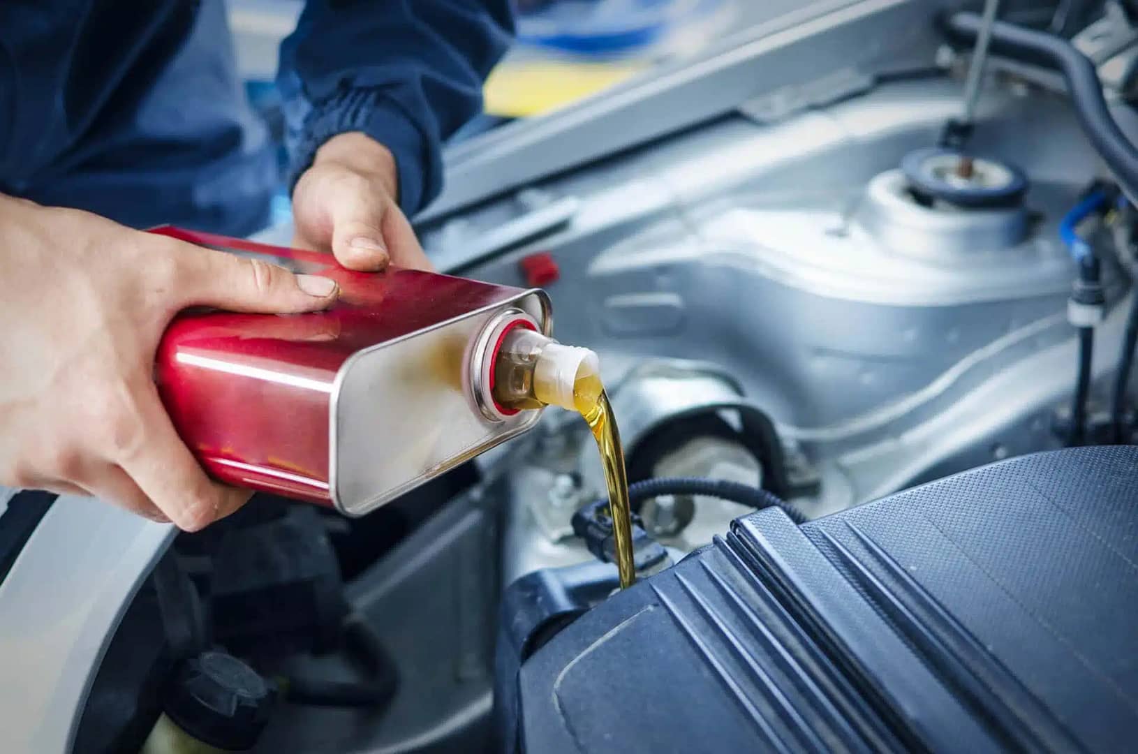 A mechanic pouring oil from a red container into a car engine, focusing on maintaining or servicing the vehicle in a garage setting.