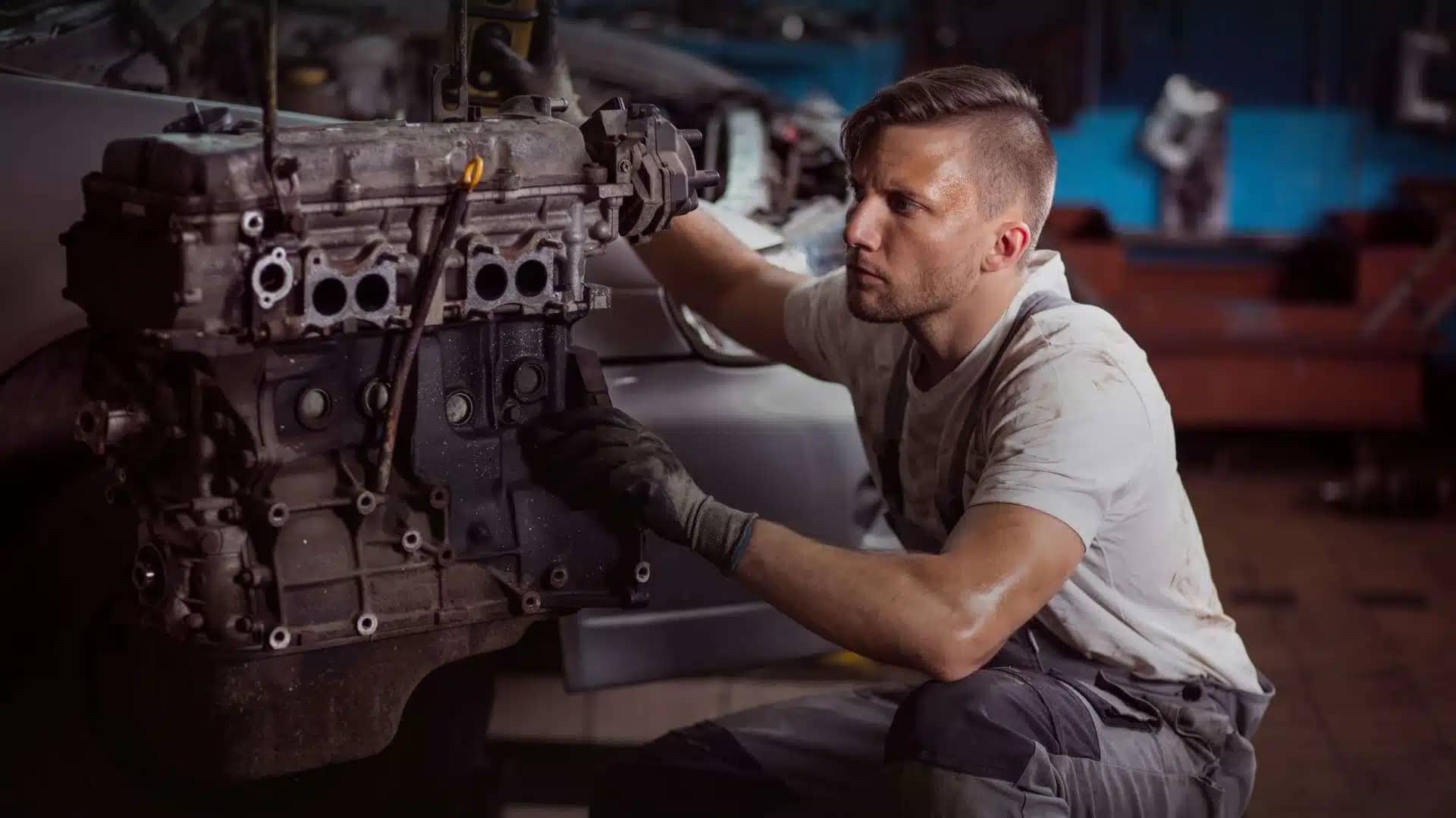 A mechanic inspects a large car engine in a workshop, holding a tool in one hand and focusing intently on his task. the garage is dimly lit with other vehicles in the background.